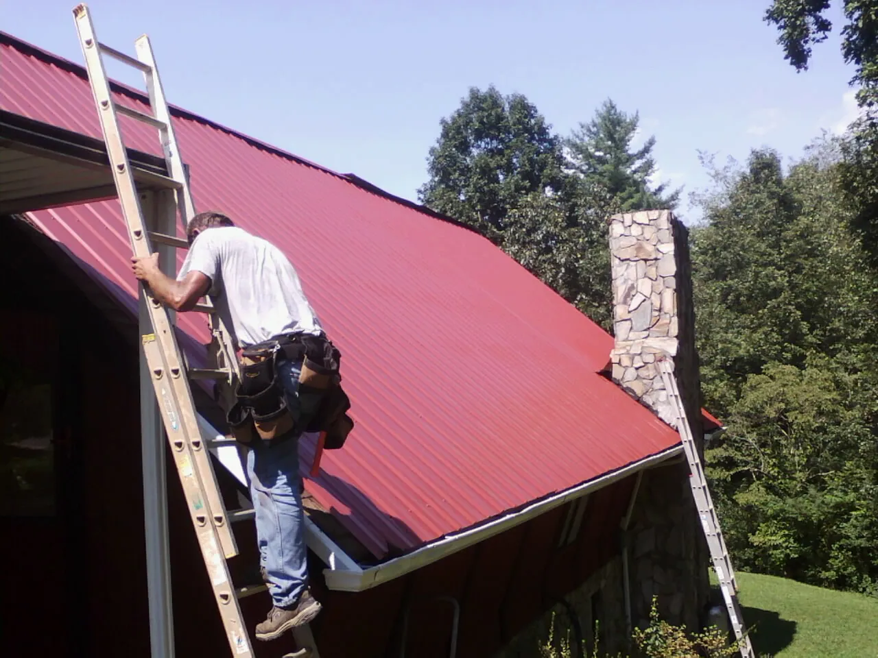 Roofer on a ladder beside a red metal roof and stone chimney — Soffit & Fascia Repair by Roofing Brothers in Fremont Center
